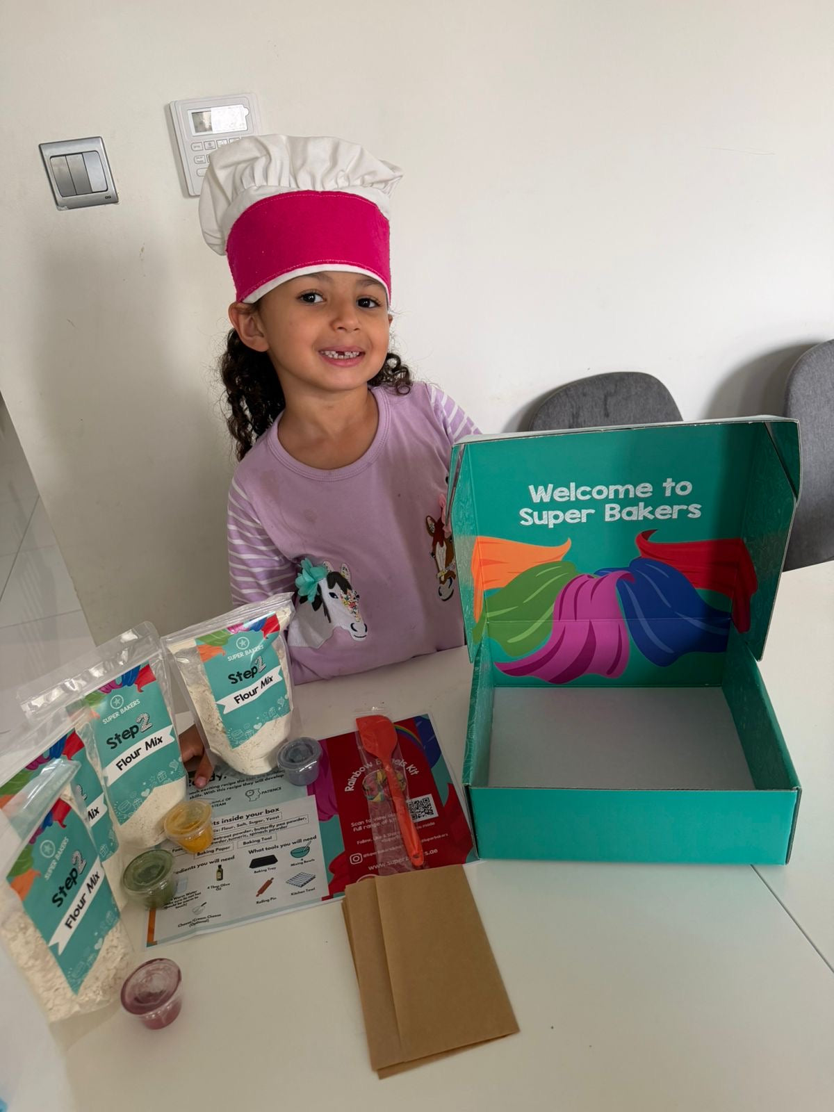 Child with a 'Super Bakers' kit on a table, including a book and baking ingredients.
